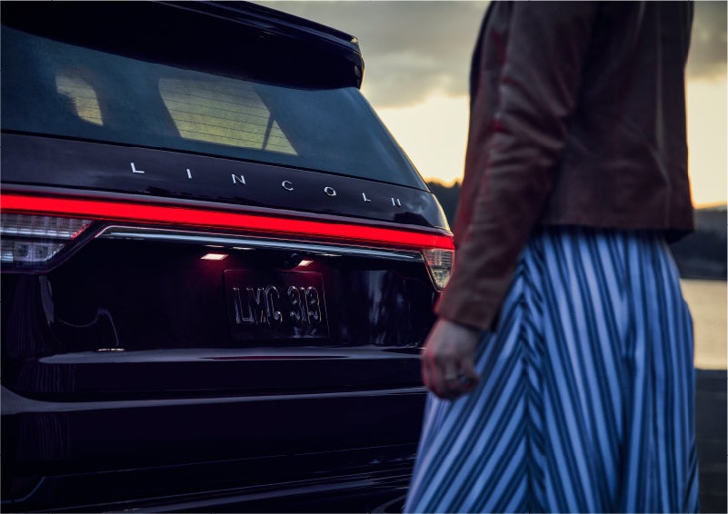 A person is shown near the rear of a 2023 Lincoln Aviator® SUV as the Lincoln Embrace illuminates the rear lights | Bayway Lincoln in Houston TX