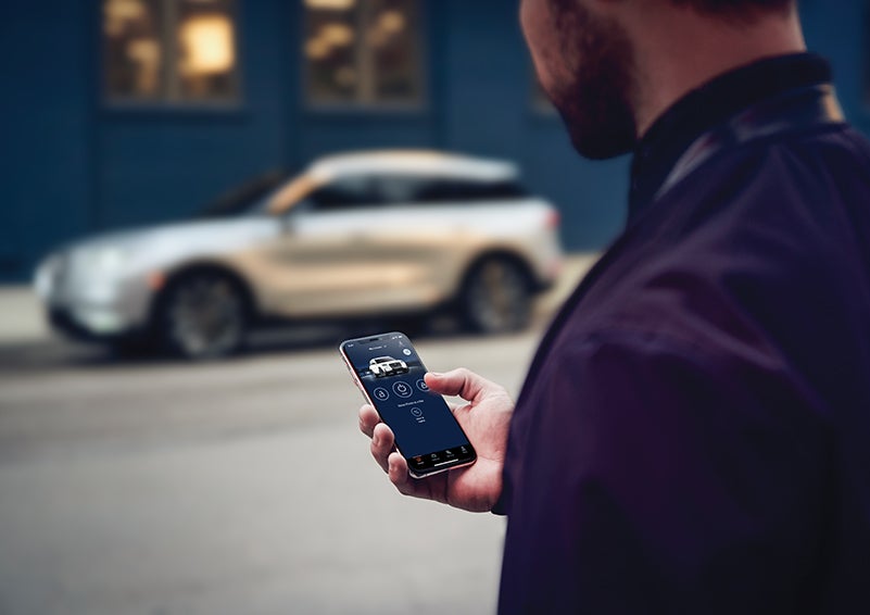 A person is shown interacting with a smartphone to connect to a Lincoln vehicle across the street. | Bayway Lincoln in Houston TX