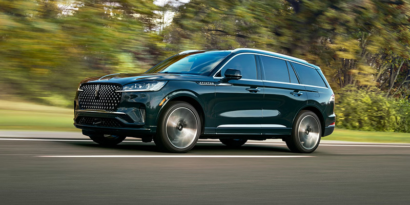 A 2026 Lincoln Aviator SUV drives along a road in Houston, Texas, with motion blur in the surrounding greenery indicating speed.